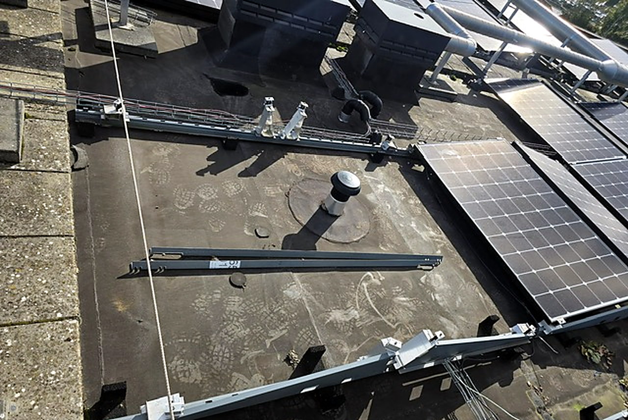 Plat dak tijdens werkzaamheden aan een zonnepaneleninstallatie: losse montagerails op rubberen beschermvoeten, een ontluchtingspijp met ronde kap en rijen gemonteerde zonnepanelen aan de rechterkant
